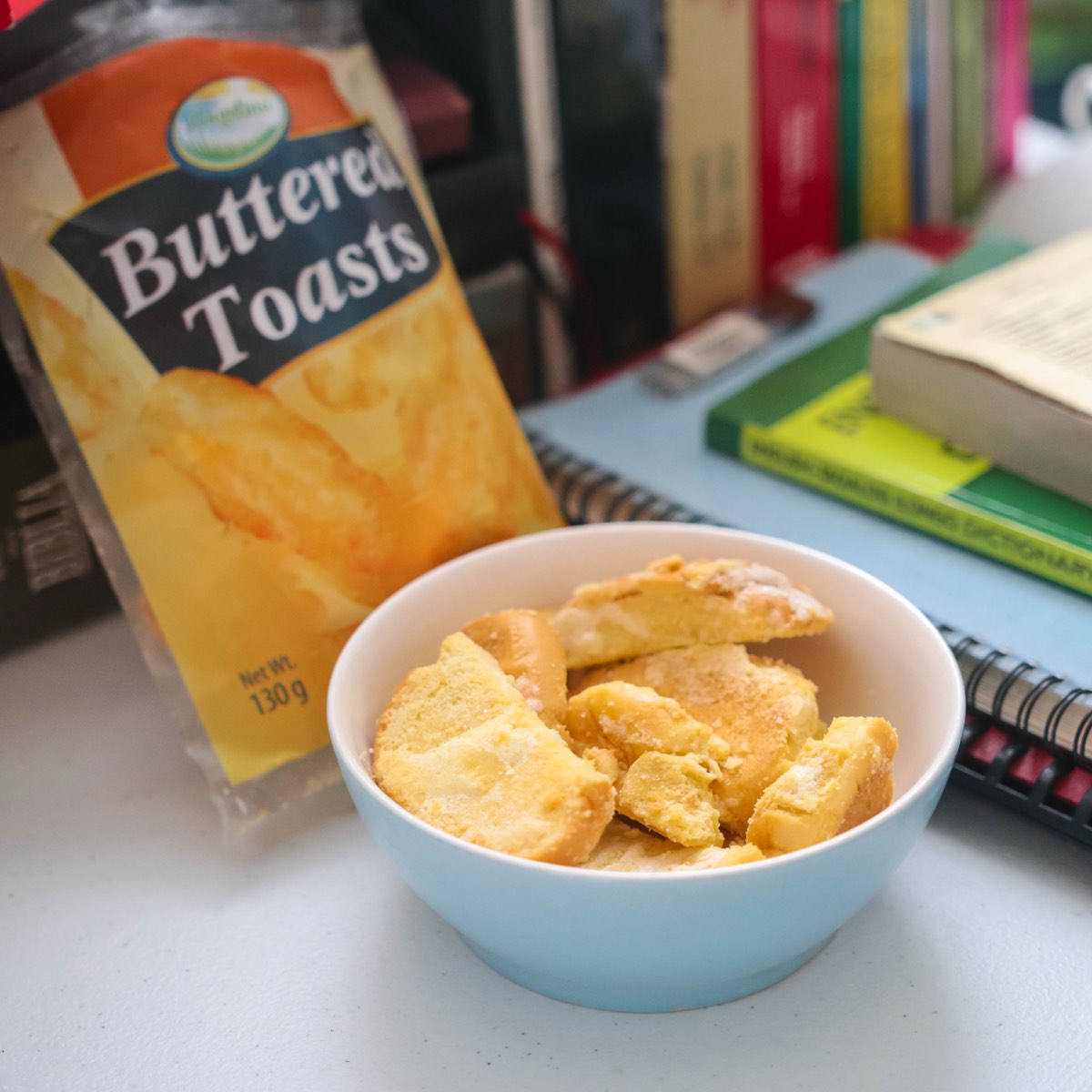 A bowl of Angelina Buttered Toasts beside the pack, styled on a desk with notebooks and a stack of books behind it.