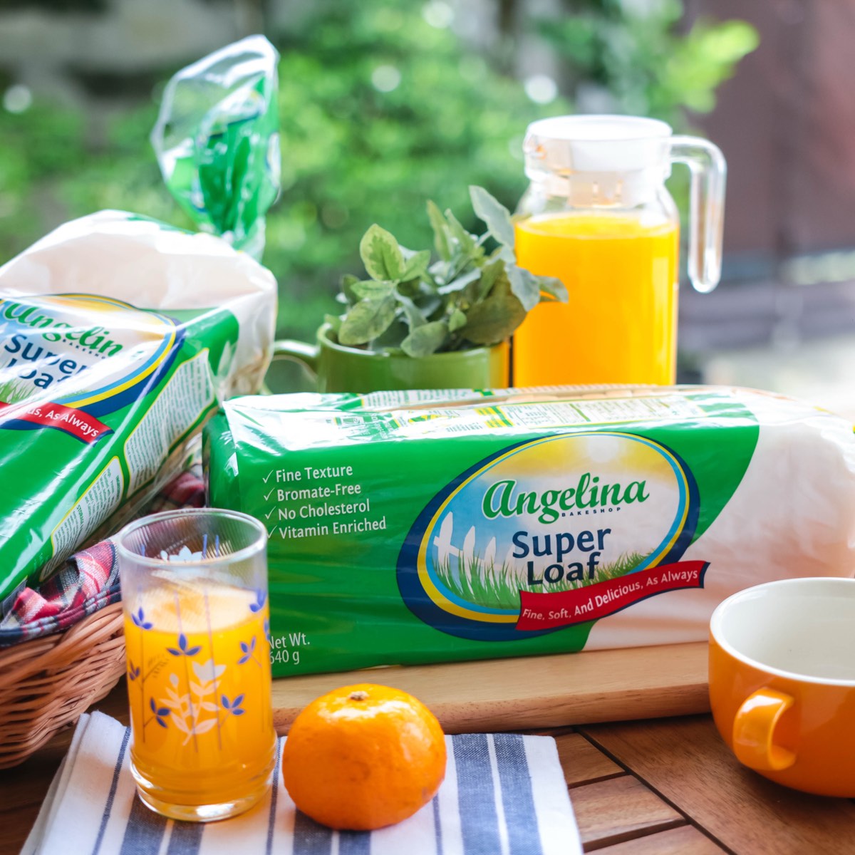 An Angelina Super Loaf on a wooden board outdoors, styled with a basket, a glass and pitcher of orange juice, a mandarin and a mug.