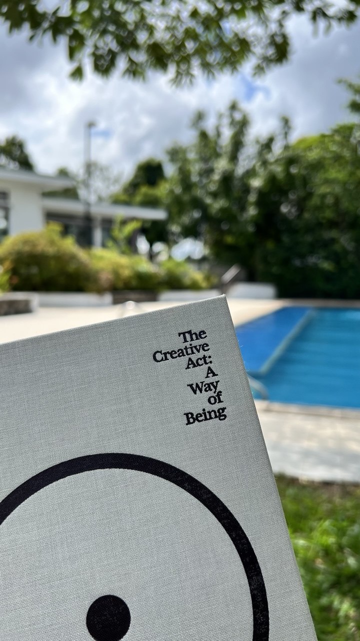 The cover of The Creative Act: A Way of Being by Rick Rubin, held up outdoors in front of a swimming pool and trees.